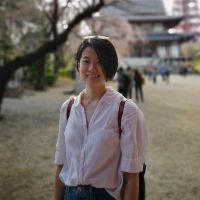 Di standing in front a sakura tree, wearing a white shirt and a smile. She is a 30-year old Chinese woman, with short black hair and no glasses in this photo.